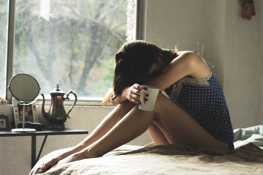 Woman sitting on bed holding a mug of coffee with head between her knees to depict how to survive an affair.