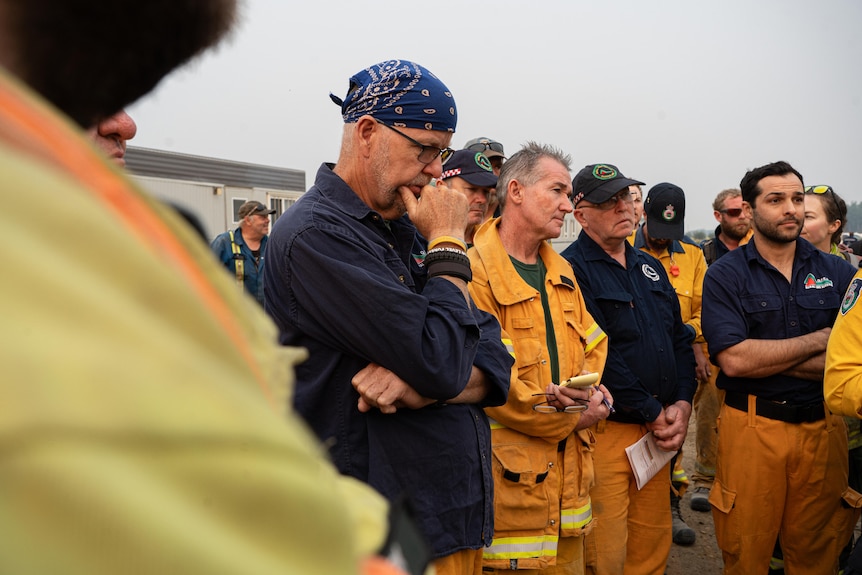 Men in firefighting outfits stand around and listen to a morning briefing. One rests his hand on his chin, contemplative.