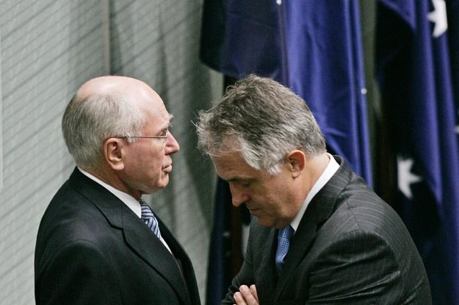 A young Malcolm Turnbull bows his head and folds his arms, standing very close to John Howard.