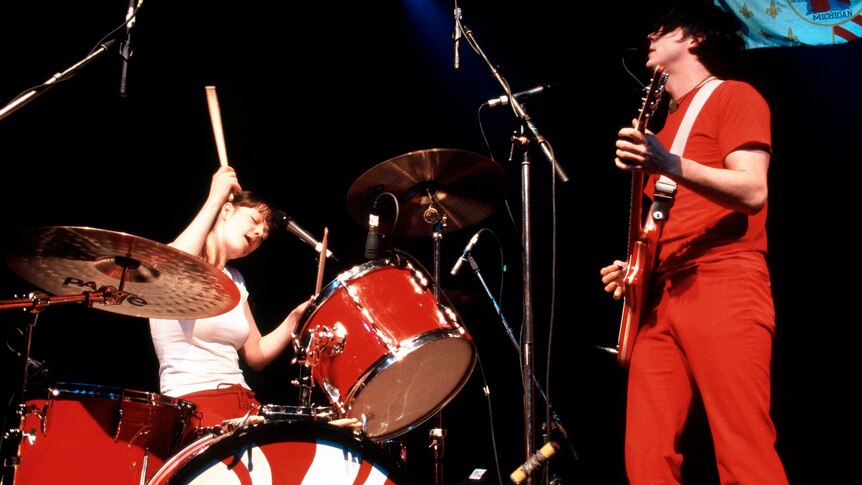 Meg White in a white t-shirt playing a red drum kit next to Jack White wearing all red playing a red guitar