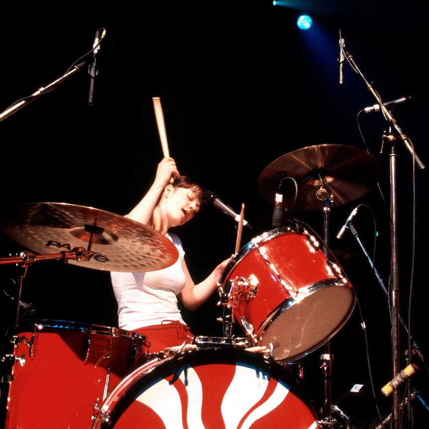 Meg White in a white t-shirt playing a red drum kit next to Jack White wearing all red playing a red guitar