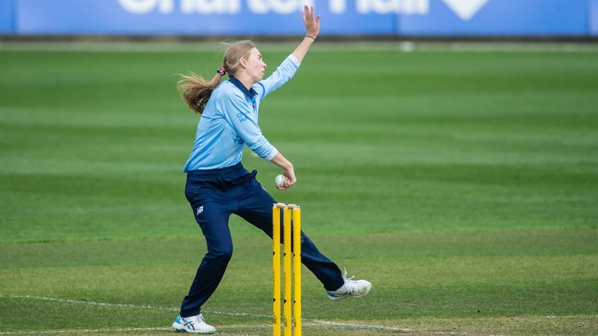 action shot of woman in blue shirt and navy pants bowls a ball next to yellow cricket stumps
