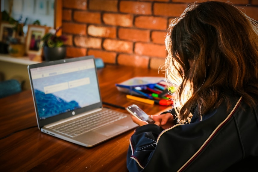 A school student at a table at home, using a learning program on a laptop and looking at an iPhone.