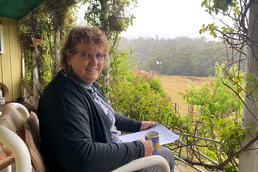 A woman sitting on a veranda overlooking farmland and bush, holding a cup of tea and a printed document.