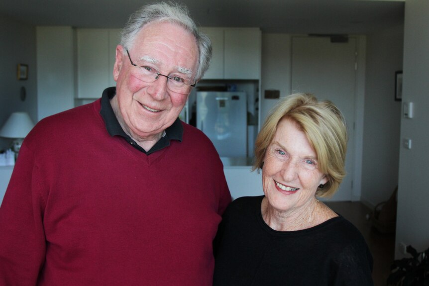 Man and woman in their late seventies smiling inside the living room and kitchen of their new apartment.