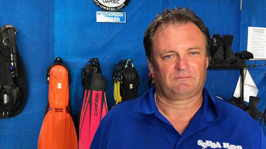 A man in a blue shirt stands in front of flippers and other scuba gear.