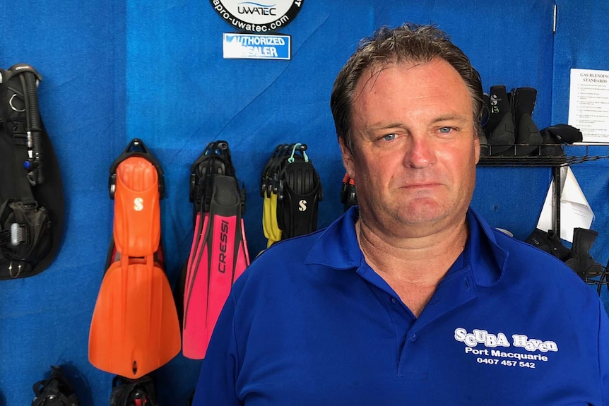 A man in a blue shirt stands in front of flippers and other scuba gear.