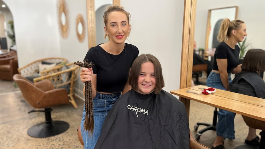 A woman in a black top and jeans holding a series of plaits standing next to a smiling young girl with short hair