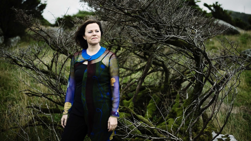 Pianist Kathryn Stott wears a blue and black full-length dress and stands in front of a tree