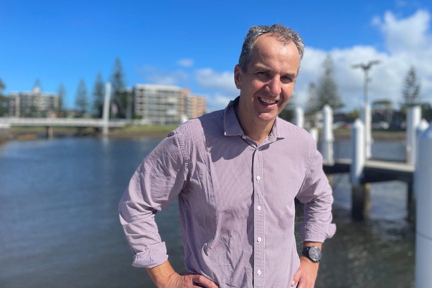 A man in a shirt and trousers stands in front a river in a coastal town, smiling.