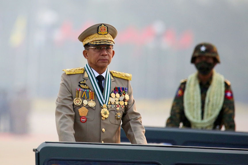 An elderly Asian man in decorative military uniform stands at a podium with a military parade behind him.