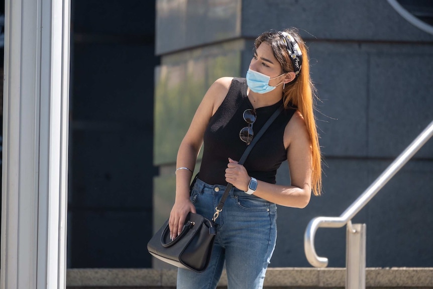 A woman with long hair wearing a face mask and black vest looks to her left.