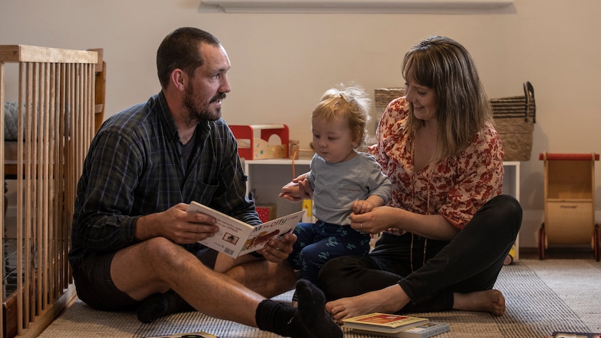 A young couple sit on the floor with their baby son and read a book Dear Zoo