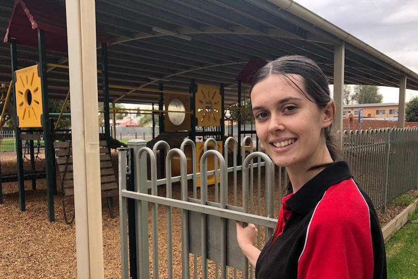 A young woman has her hand on the fence at the gate to an undercover playground.