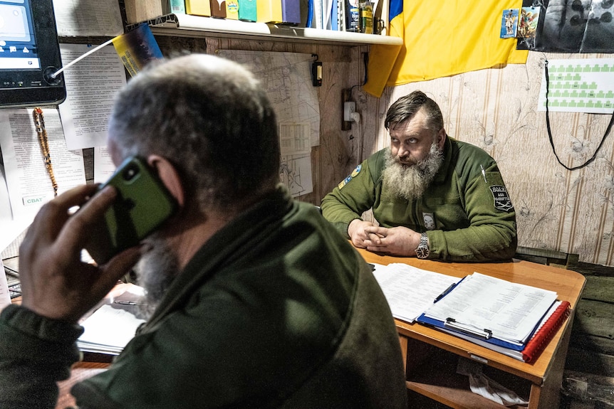 A heavily bearded man in a green military jacket looks up at a screen from where he sits behind a desk in a cramped space