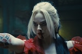 Margot Robbie as Harley Quinn has a bloody nose, is wearing a red dress, and has her arms outstretched, with a determined look