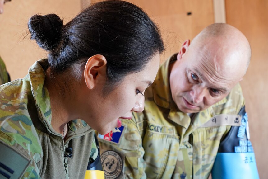 A woman with black hair tied in a bun and wearing army fatigues talks to a bald man wearing army fatigues.