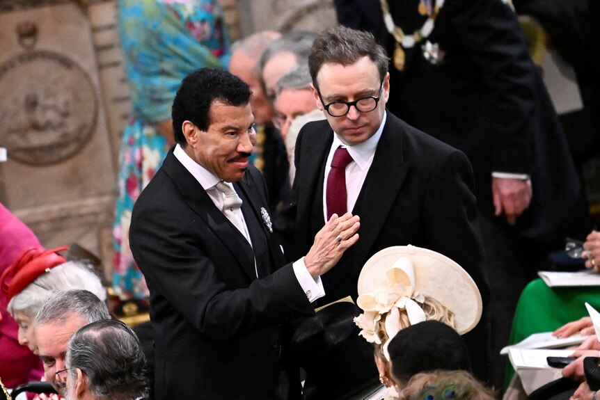 Lionel Richie in a black and white suit with a hand raised, standing in a crowd. 