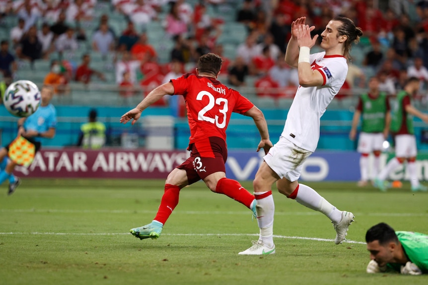 A defender raises his hands to his head as the ball goes into the net, and a striker wheels away in celebration.