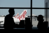 The silhouettes of two people inside a Perth Airport terminal with Virgin planes outside the window.