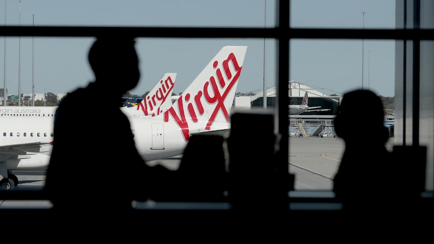 The silhouettes of two people inside a Perth Airport terminal with Virgin planes outside the window.