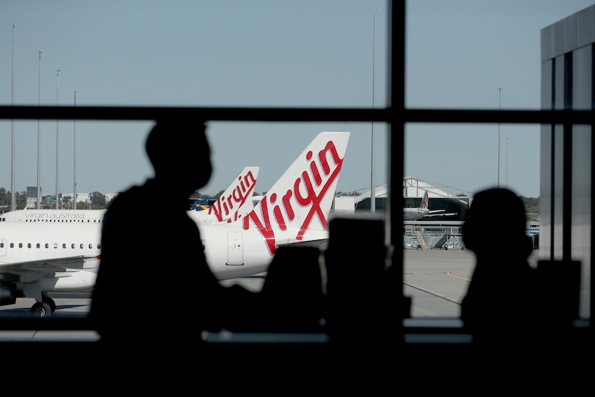 The silhouettes of two people inside a Perth Airport terminal with Virgin planes outside the window.