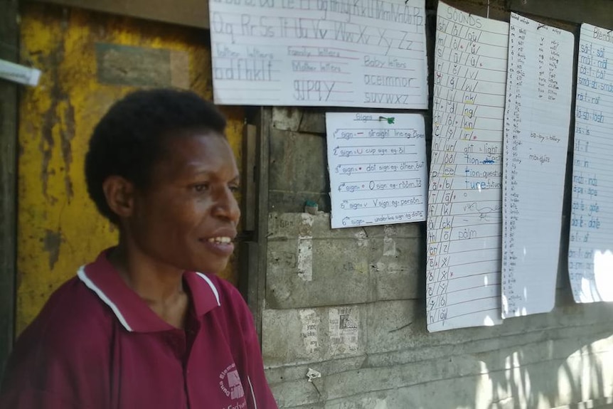 Ruthy Cletus stands in front of a makeshift blackboard made up of paper nailed to a wall with writing on them