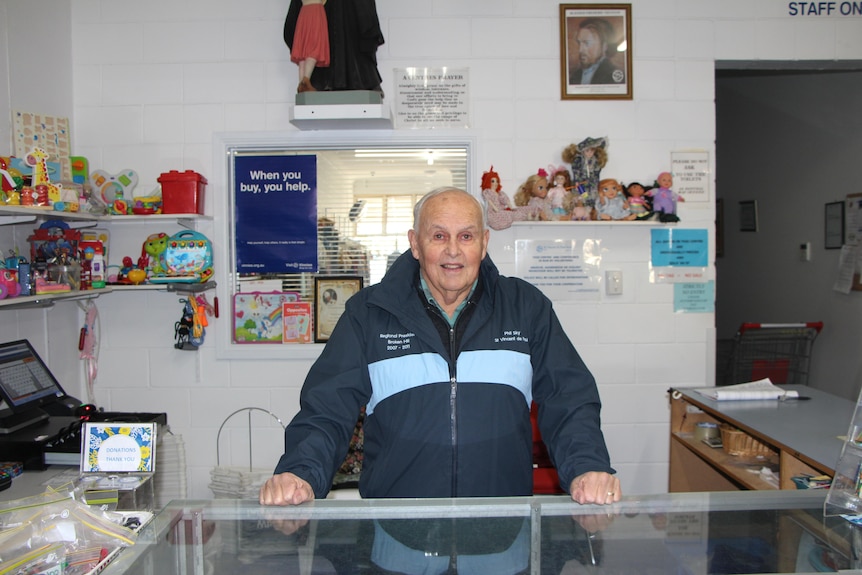 An old bald white man wearing a blue jacket standing behind a shop desk 
