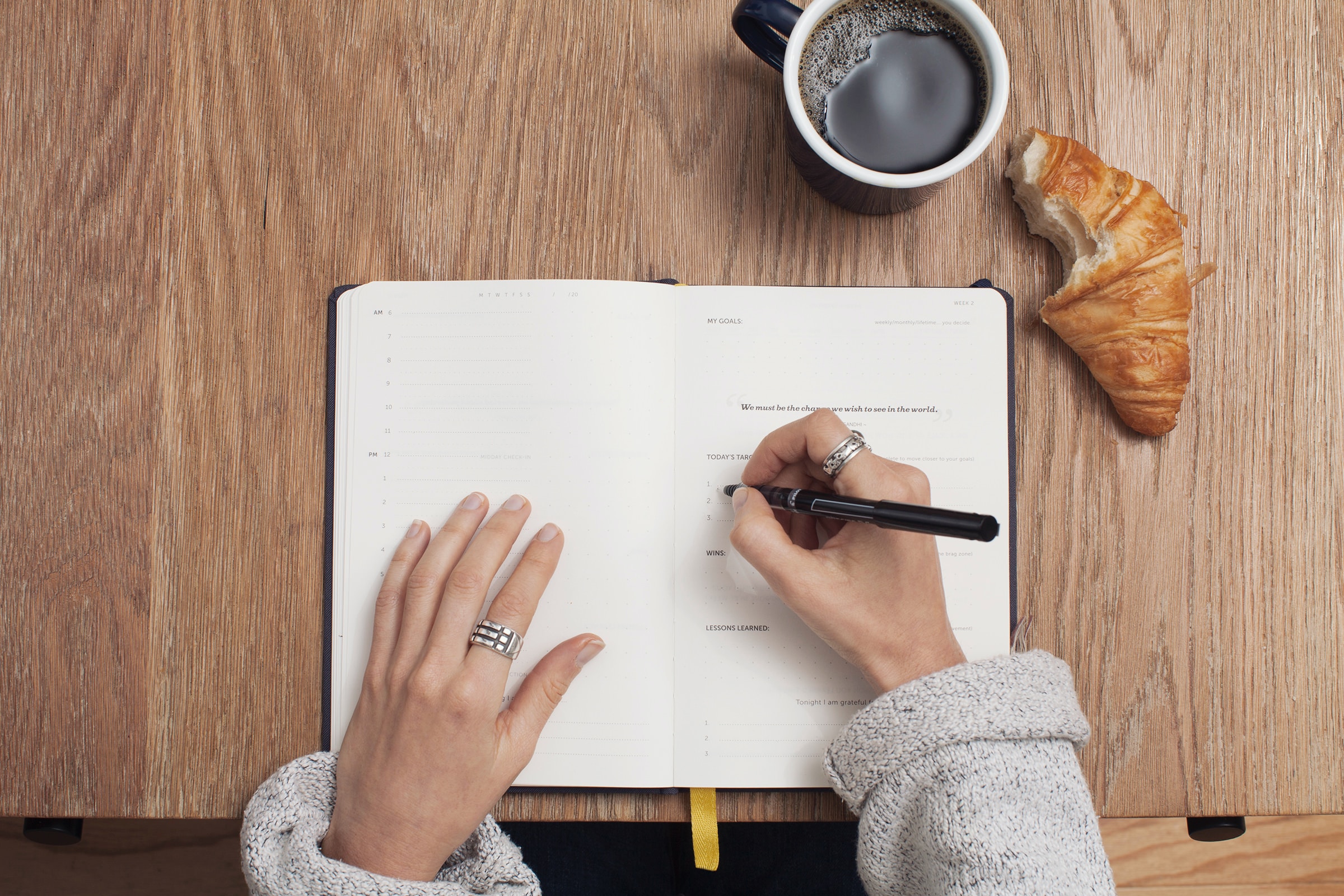 A close-up of a person's hands writing in a book on a desk, with a croissant and coffee nearby