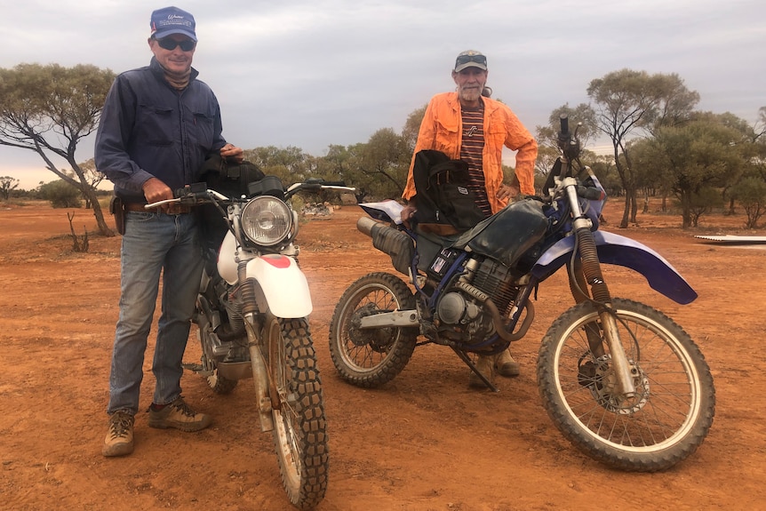 Two men stand beside two motorbikes on a red, sandy plain.