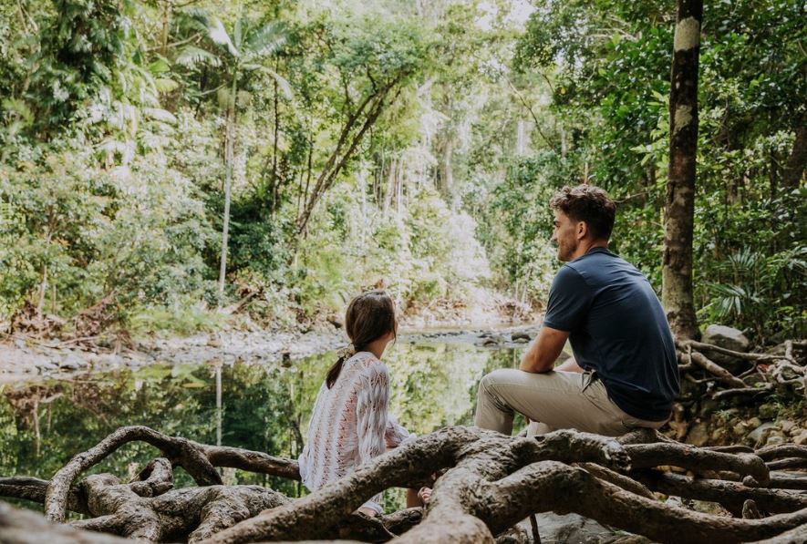 A man and a woman sitting on a clump of tree roots, looking at a stream in an ancient rainforest.
