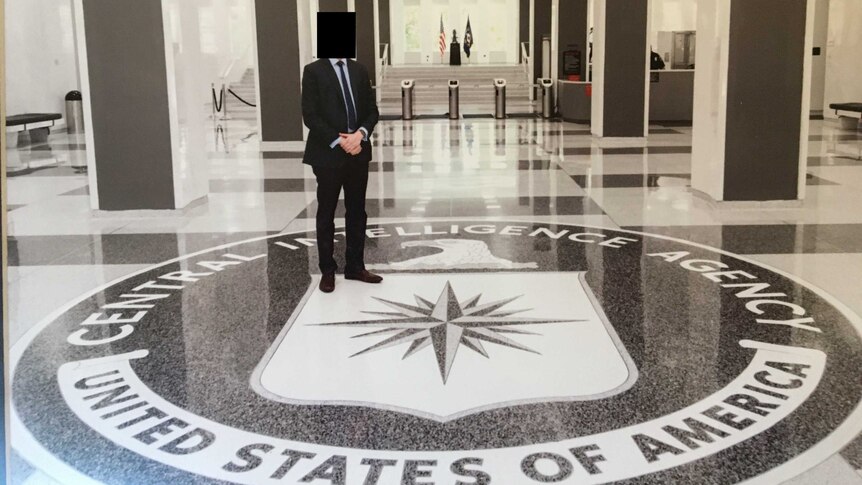 A man in a suit with a blurred out face stands in a foyer above a floor plaque for the CIA