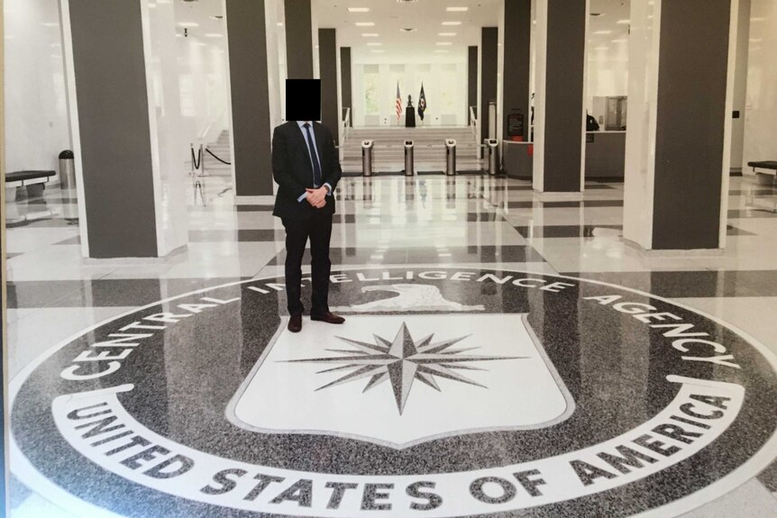 Un homme en costume avec un visage flou se tient dans un hall au-dessus d'une plaque au sol pour la CIA