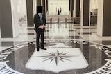 A man in a suit with a blurred out face stands in a foyer above a floor plaque for the CIA