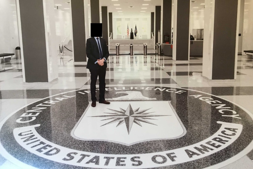 A man in a suit with a blurred out face stands in a foyer above a floor plaque for the CIA