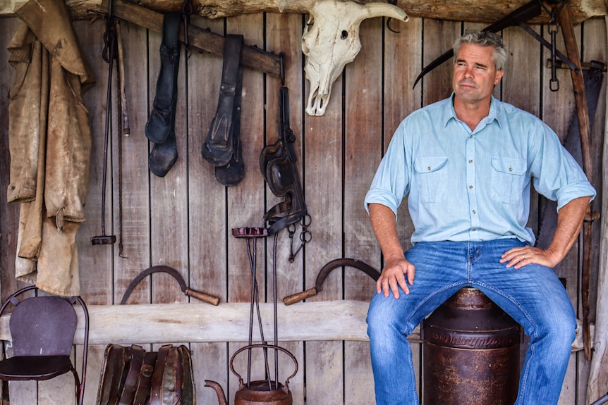 A man in a light shirt and blue jeans sits on a drum in a wooden shed decorated with old farm equipment.