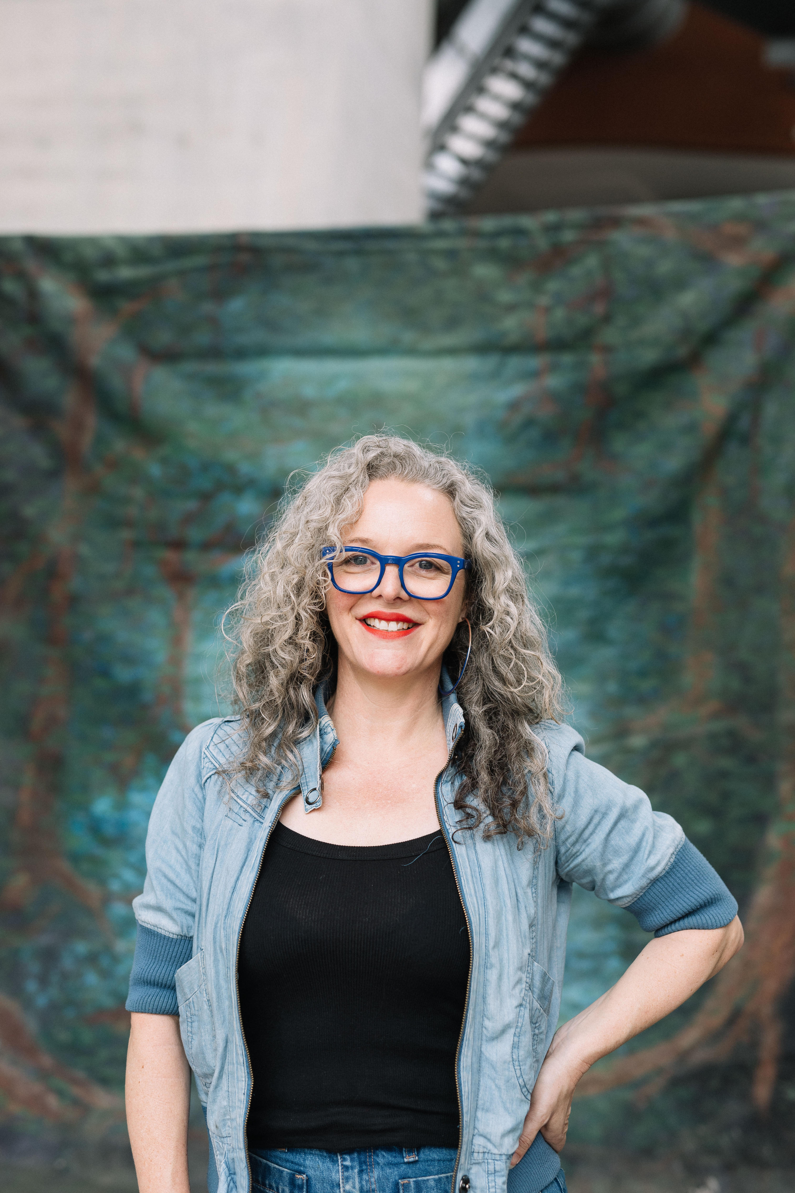 White woman with curly grey hair wears vibrant blue glasses, red lipstick and a denim jacket over a black shirt.