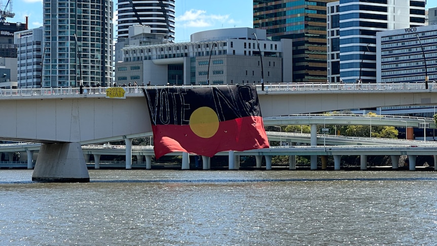 A large Aboriginal flag hanging from Brisbane's Victoria Bridge over the river with the words "vote no" written across it
