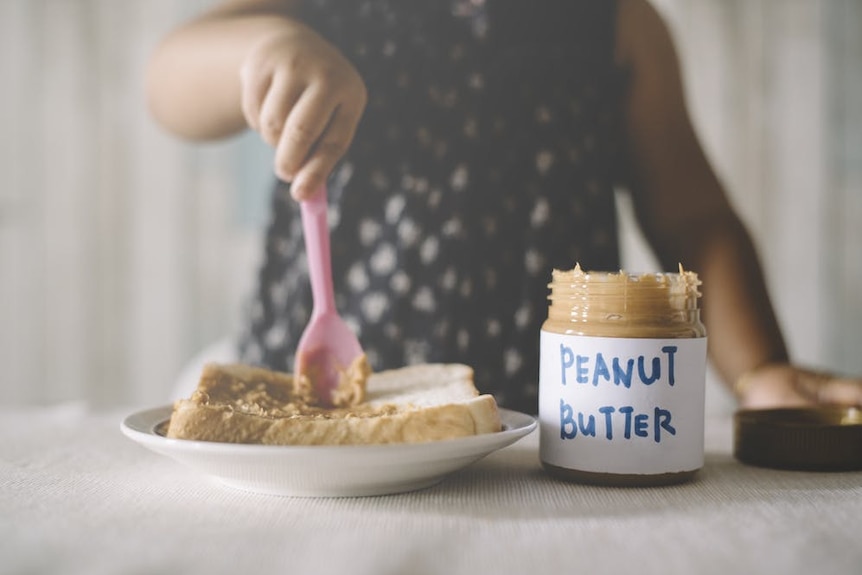 A child holds a pink spoon above a piece of bread next to a jar of butter.