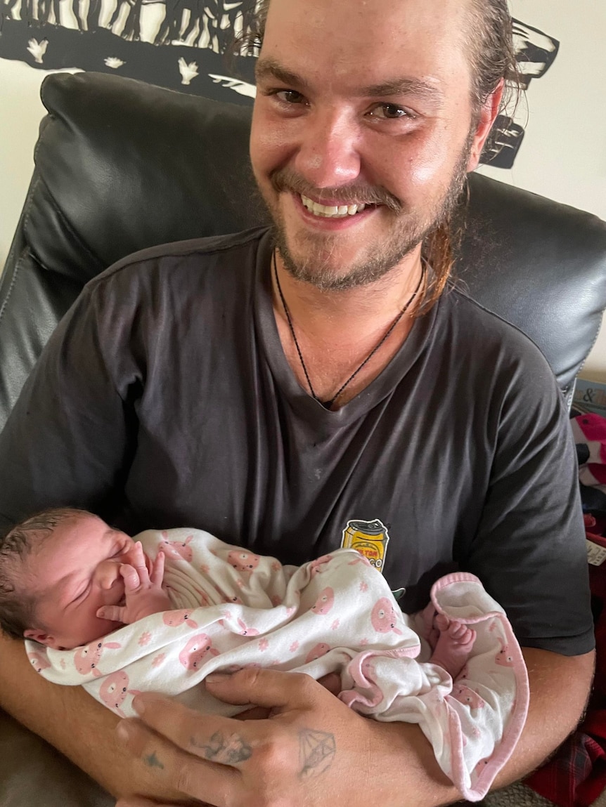 A young man smiling at the camera, holding his newborn daughter.