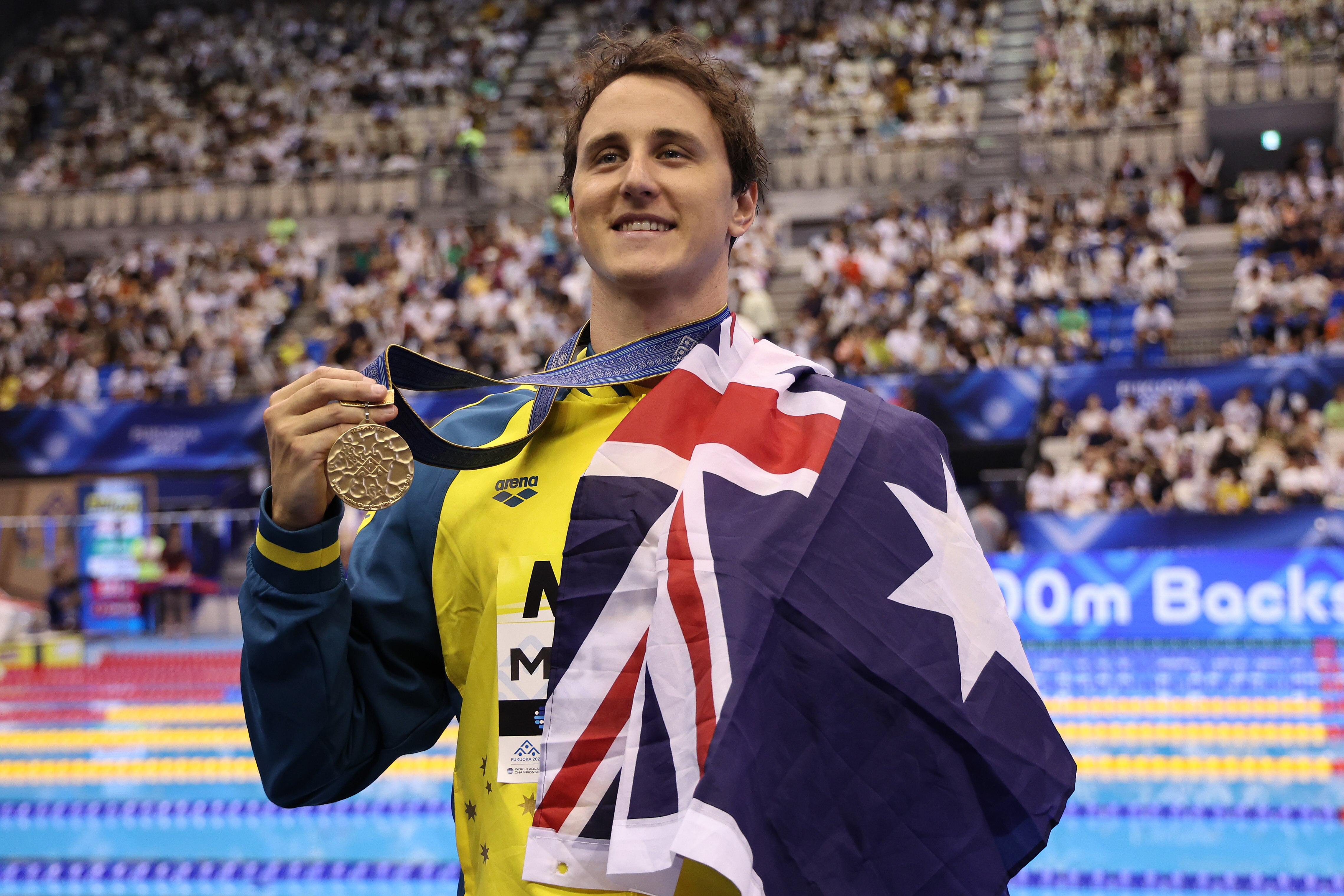 Smiling Cam McEvoy holds a gold medal, draped in Australian flag, crowds behind him.