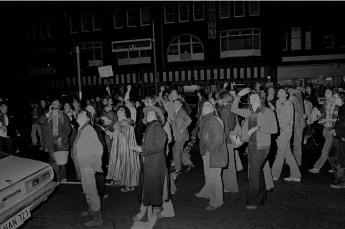 A large crowd of people on the street looking up, some waving. Black and white photo.