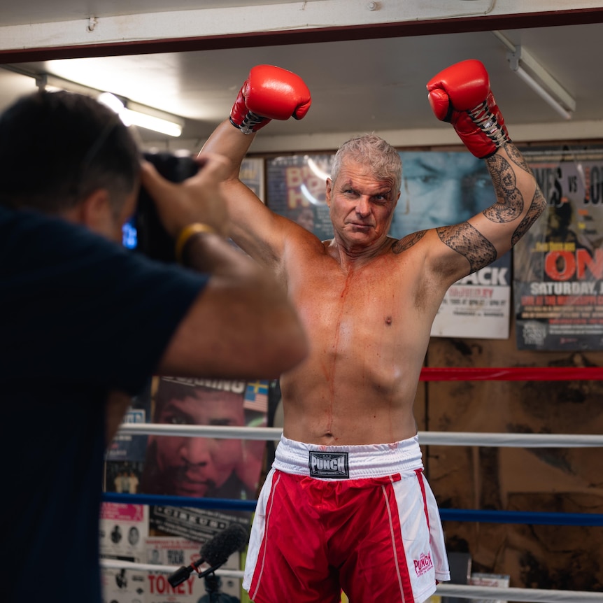 McBride shirtless and wearing boxing gloves holds his arms up in front of a photographer in a boxing ring.