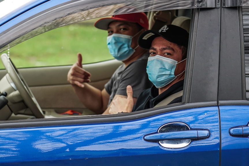 Two men in face masks sit in a car and give the thumbs up. 