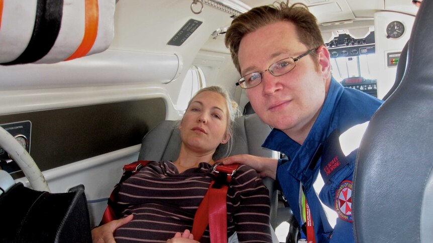 A man in a medical uniform in  an aircraft with a pregnant woman lying back on a seat.