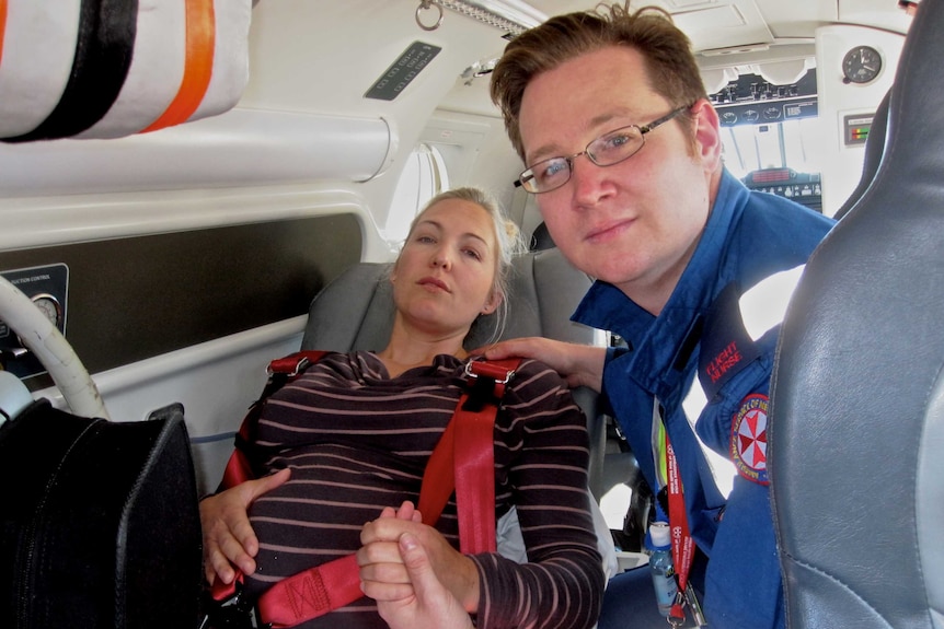 A man in a medical uniform in  an aircraft with a pregnant woman lying back on a seat.