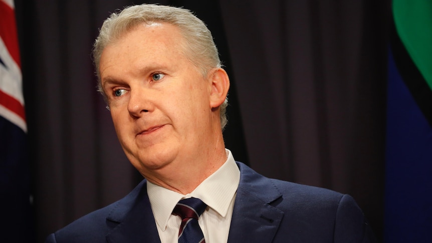 A middle-aged white man with grey hair in a suit stands in front of a blue curtain.