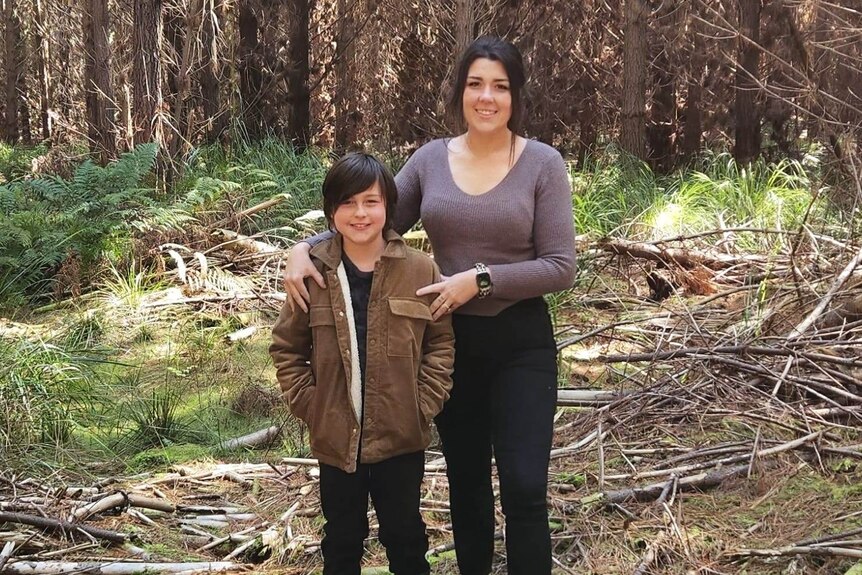 A smiling dark haired woman stands next to a boy with her arm around him 