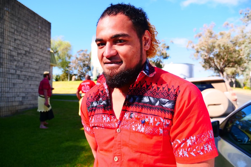 A Tongan man stands in a red shirt.