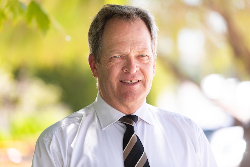 A man in a white shirt with a black and gold tie smiles into the camera.